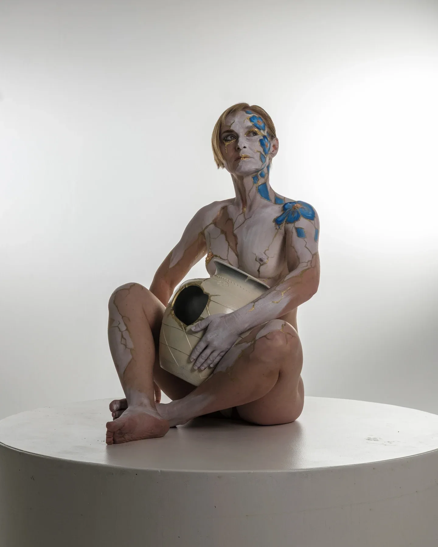 Tiana sits on a white platform holding a pale vessel with a dark opening; blue and gold body paint crosses her face and arms.