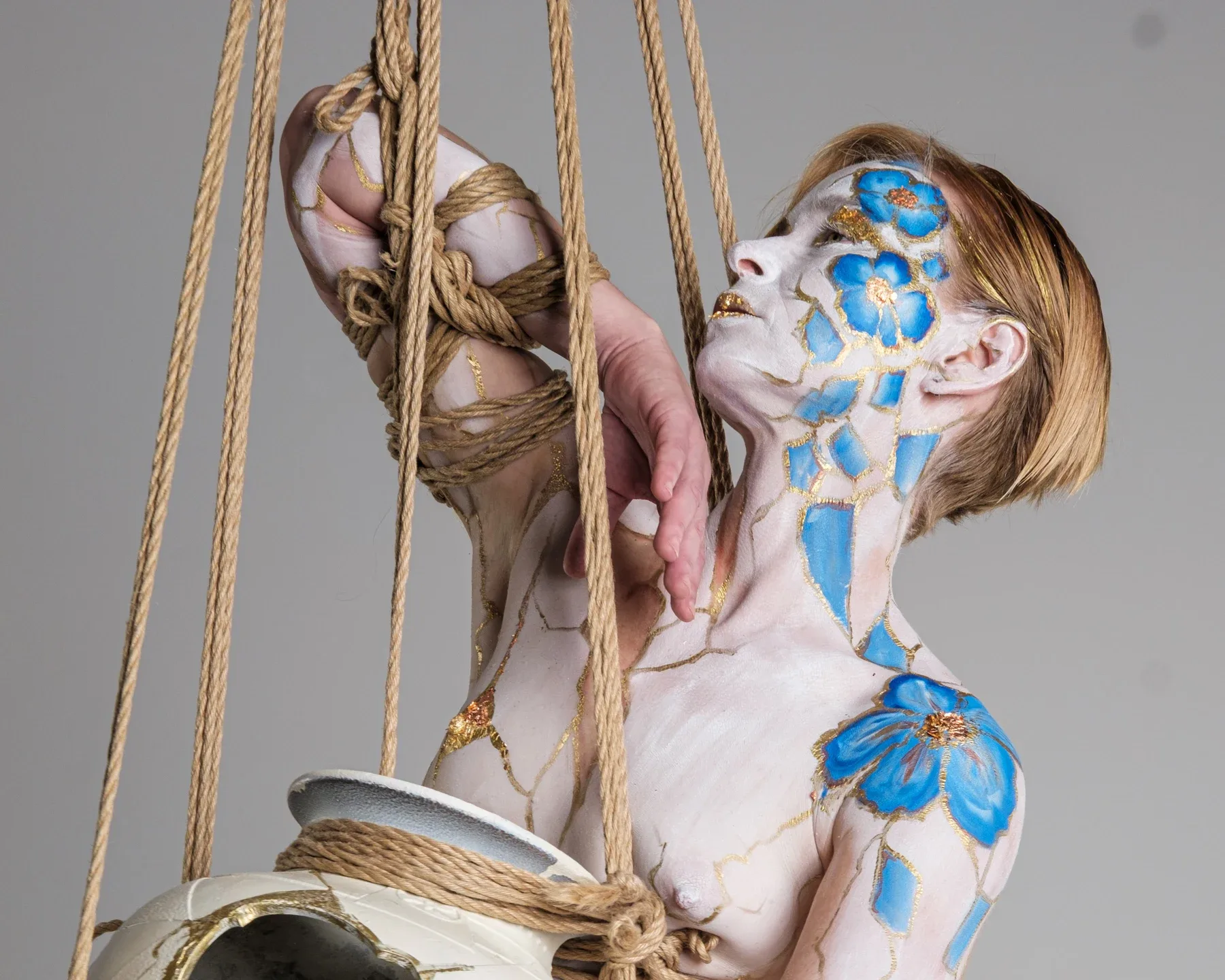 Close view of Tiana beside hanging ropes, her bound hands above a pale vessel and blue flowers painted across her face and shoulder.