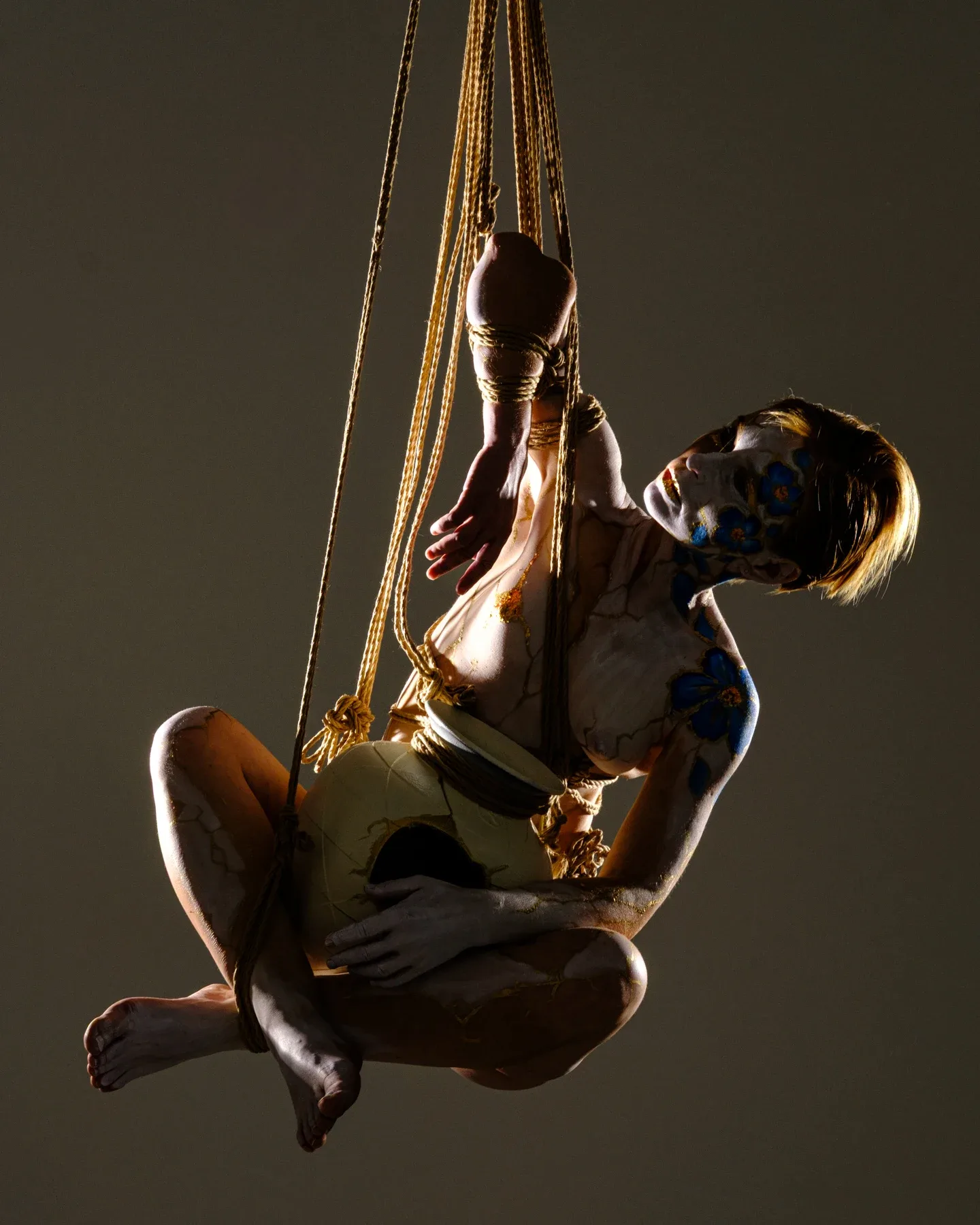 A side-lit rope suspension against a dark background: Tiana curls around the pale vessel while her face and one arm emerge from shadow.