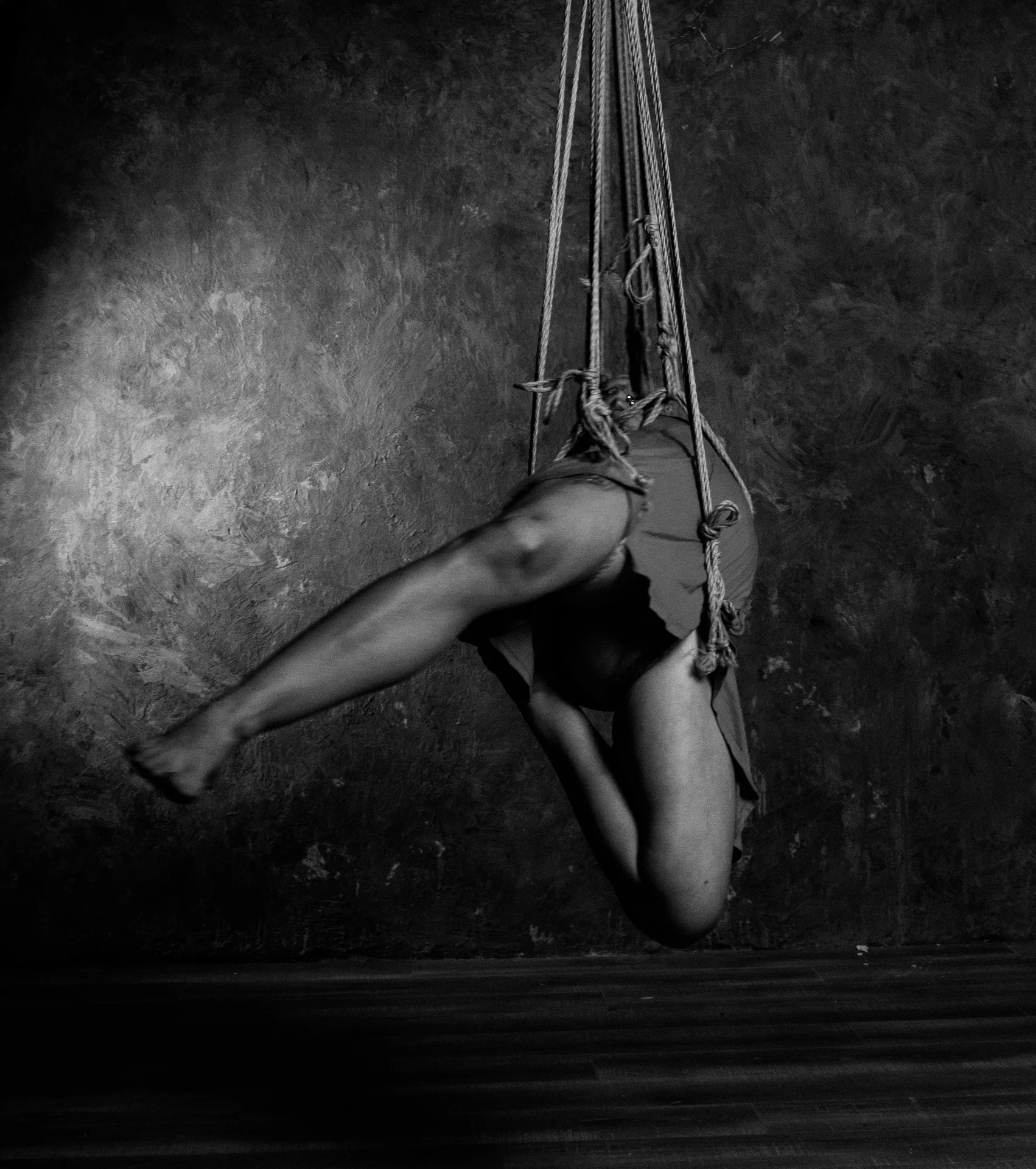 A diagonal of legs and ropes by a dark wall; the head is hidden behind the body.
