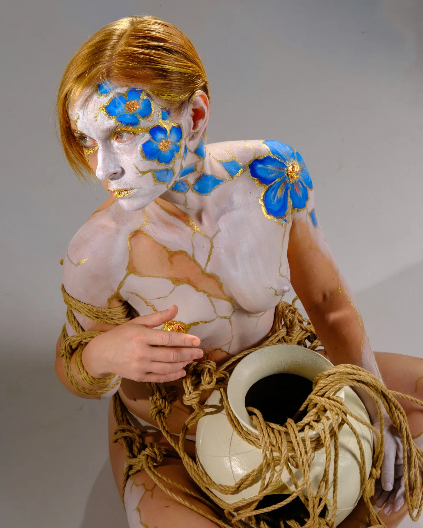 Tiana sits with loose ropes around her arms and a pale vessel in her lap, looking down toward its dark opening.