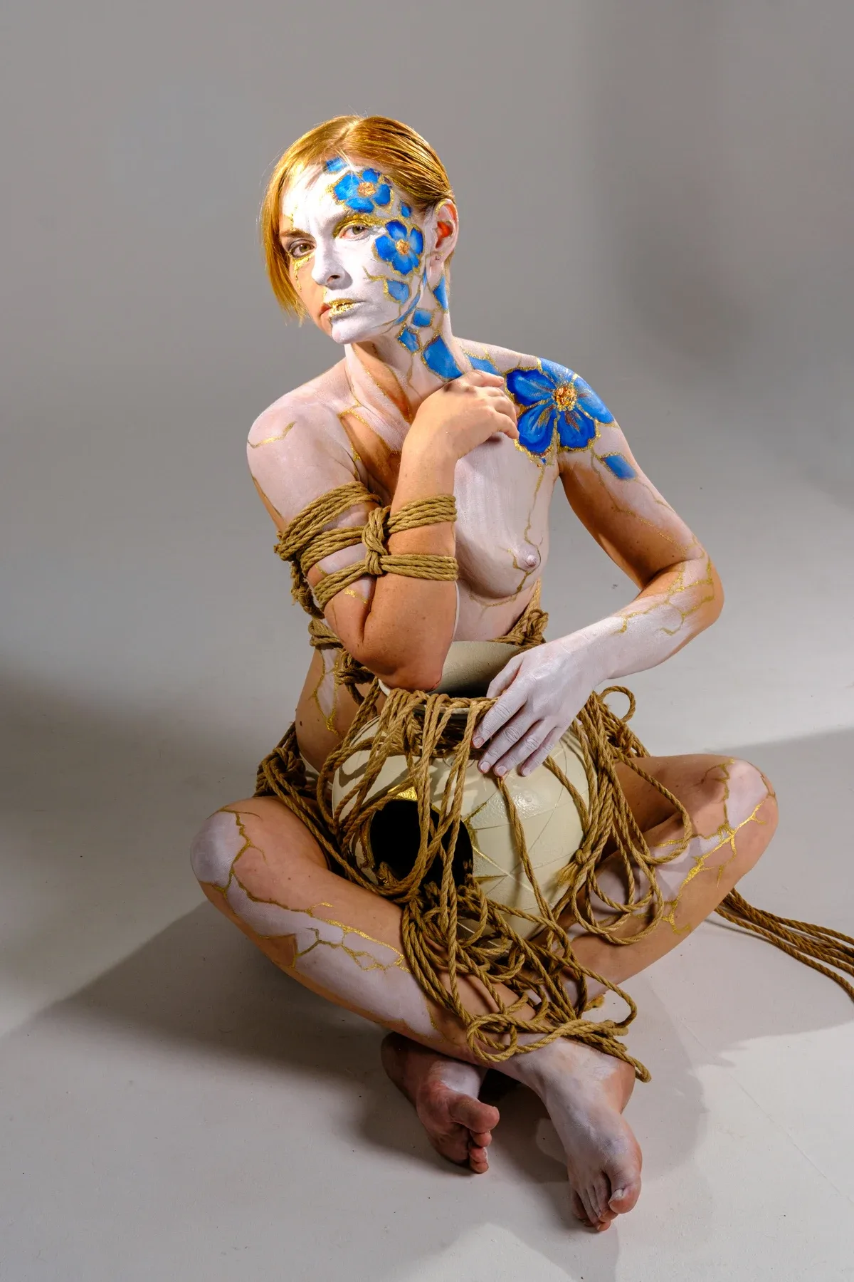 Tiana sits facing the camera with a pale vessel and a coil of rope in her lap; blue flowers and gold lines remain on her skin.