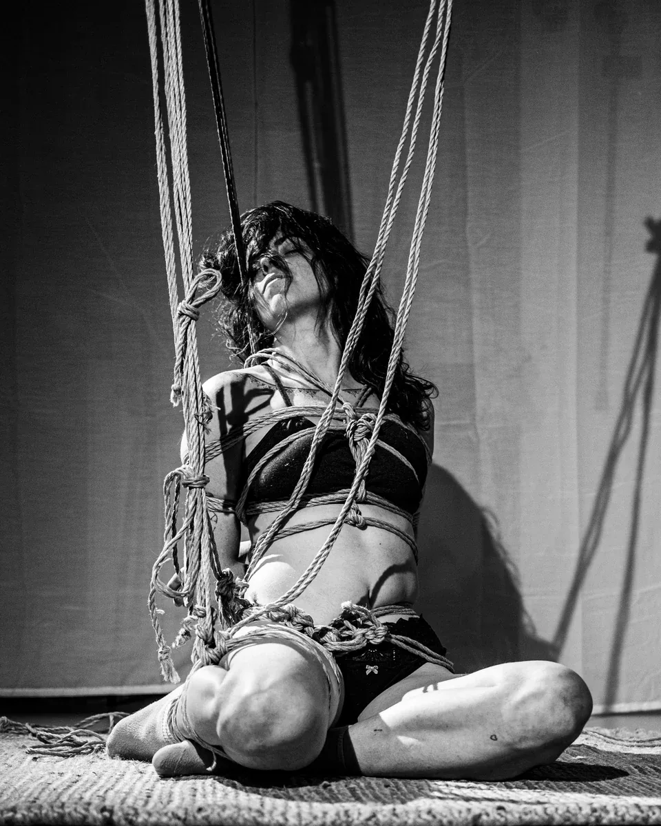 An anonymous model sits cross-legged on the floor among ropes descending vertically around her, with her head tilted back.
