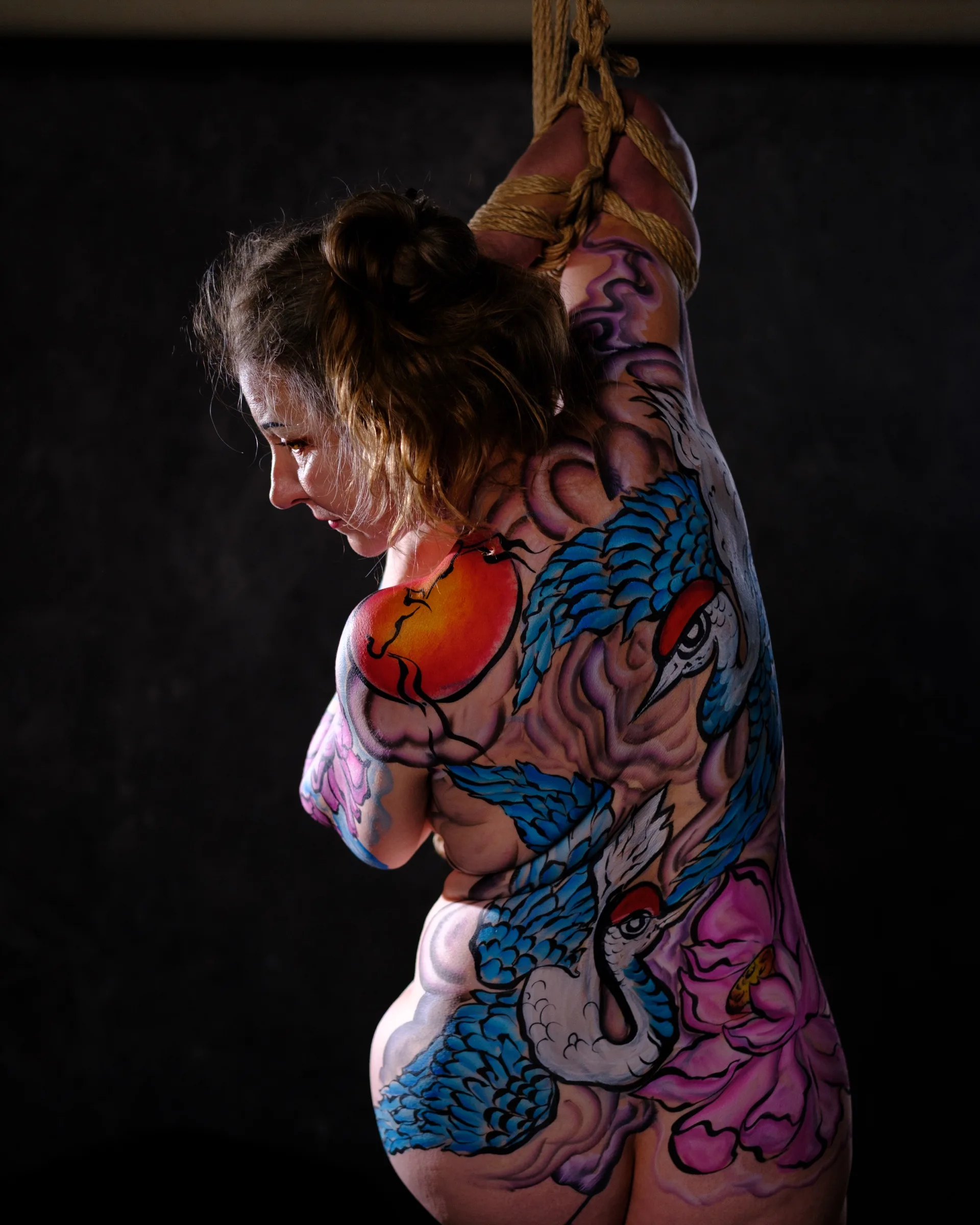 A body-painted model raises one bound arm against a dark background.