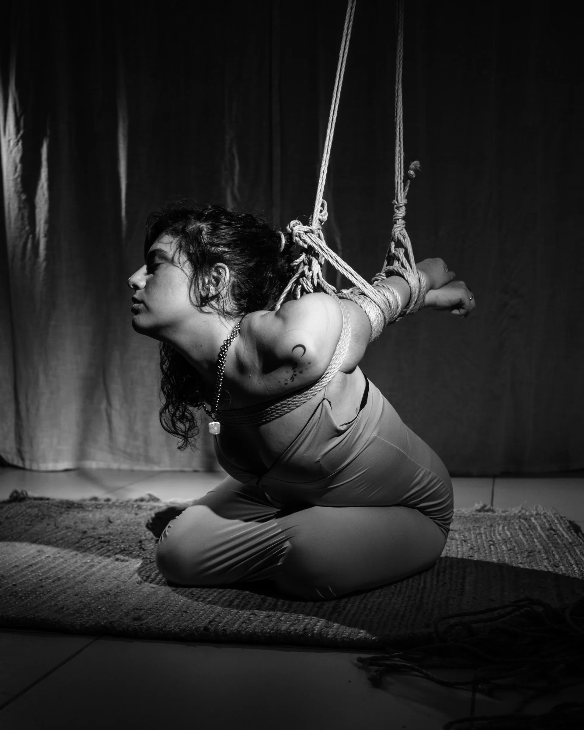 A person in a dress leans forward with their arms held behind them in rope.