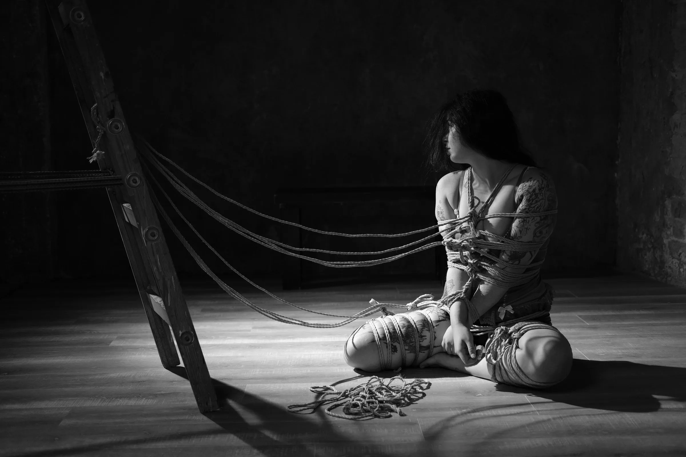 A person seated on the floor, connected by lines of rope to a leaning frame.