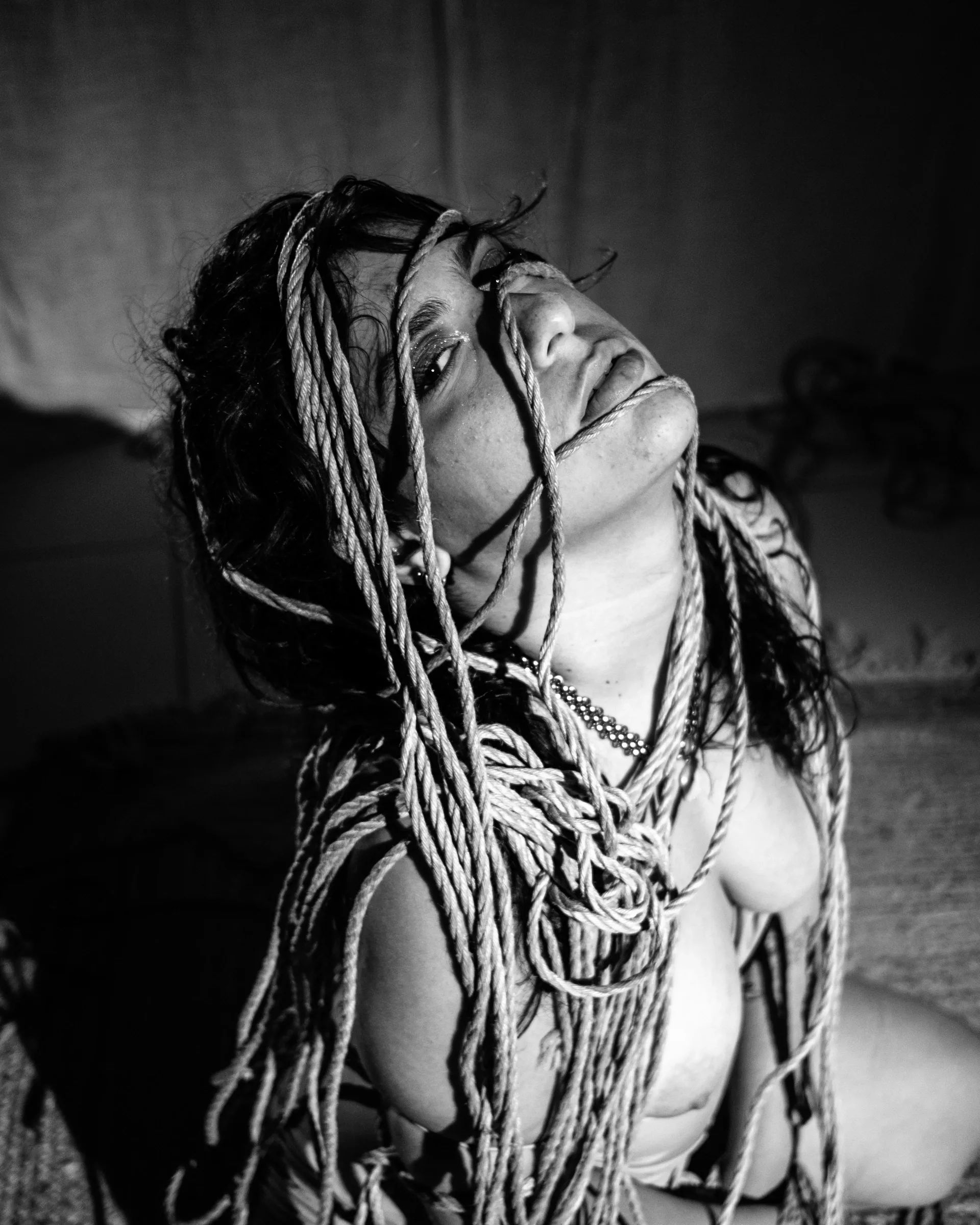 Expressive black-and-white portrait with an upturned face and ropes around the head and shoulders.