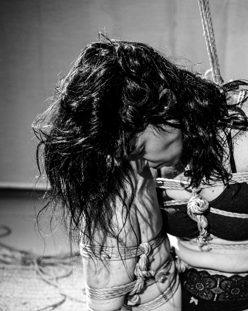 Close black-and-white portrait of an anonymous model leaning forward, dark hair covering most of her face and rope crossing her shoulder and torso.