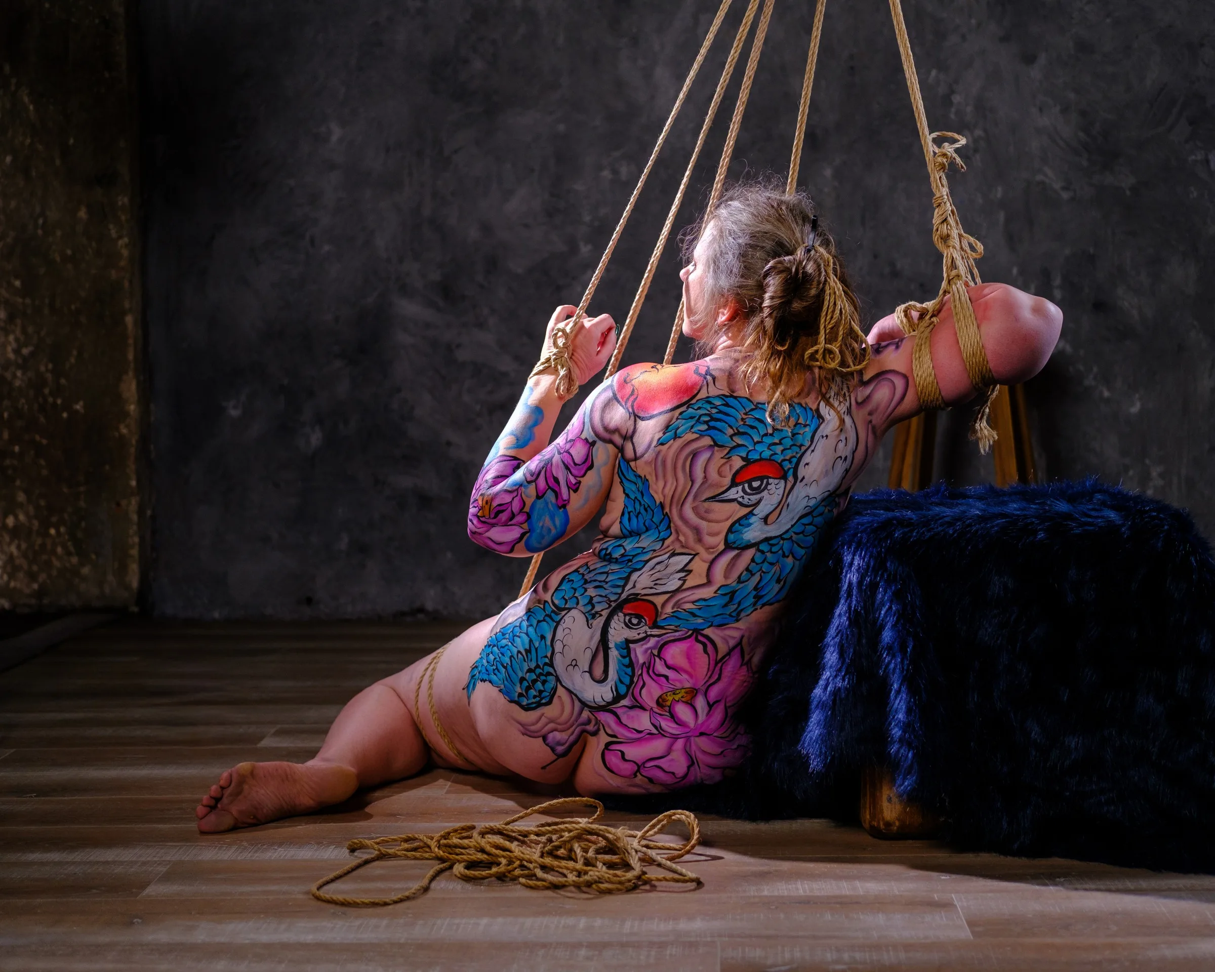 A body-painted figure sits beside a dark blue support, holding the ropes above.